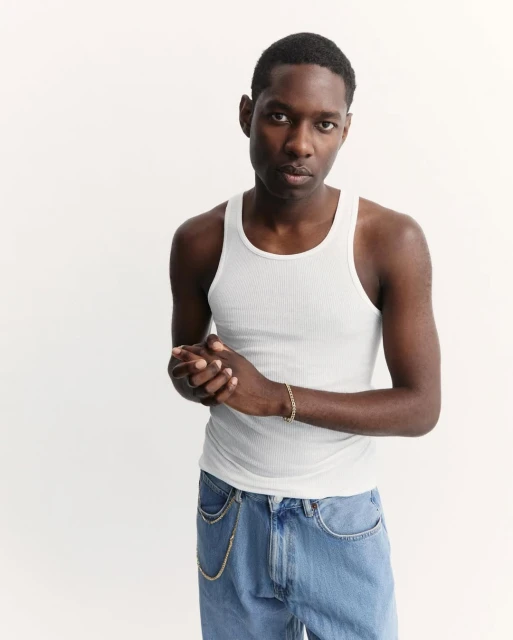 Portrait of a young Black male in a white ribbed tank top and light wash jeans, posing against a bright white backdrop in a studio setting.