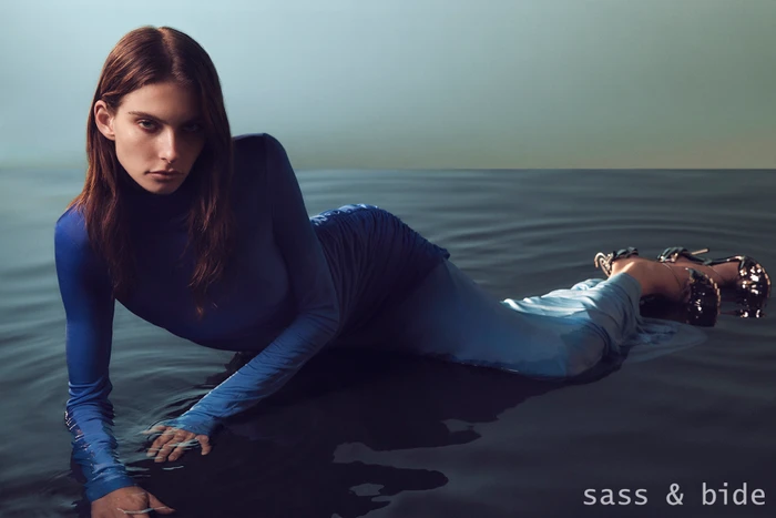 A female model wearing a blue long-sleeve top lies in shallow water, posed against a dark moody background.