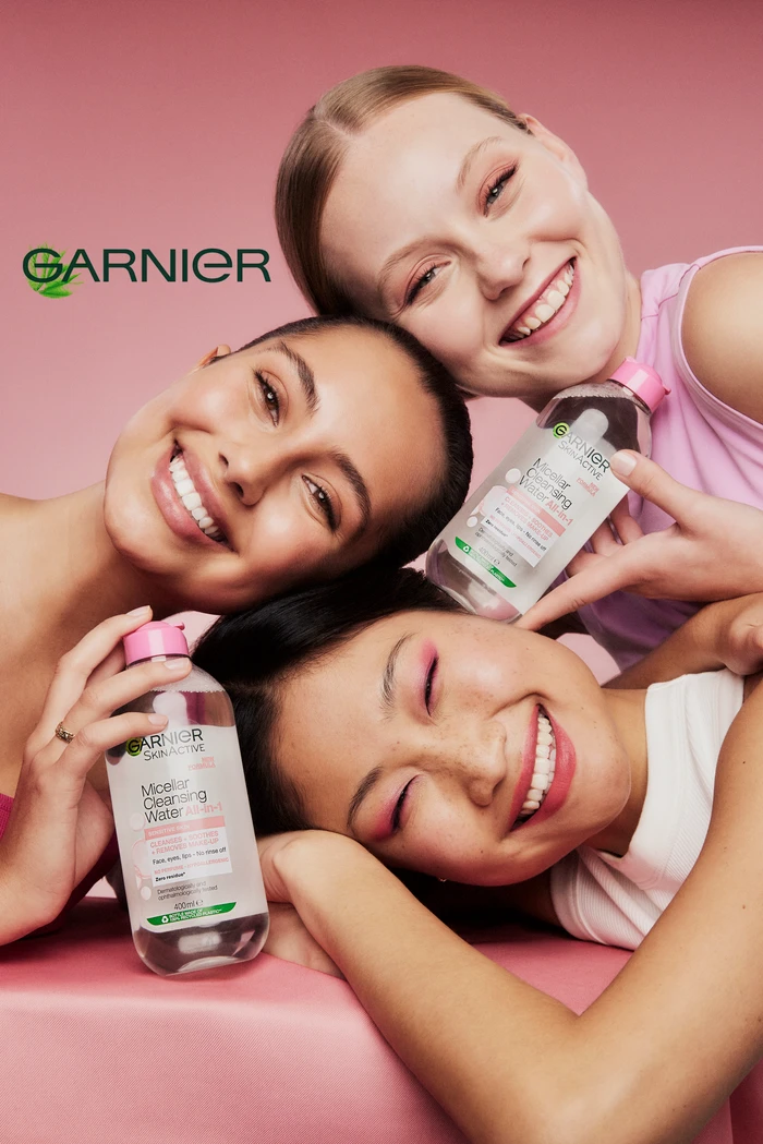 Three smiling women holding bottles of Garnier micellar cleansing water against a solid pink backdrop
