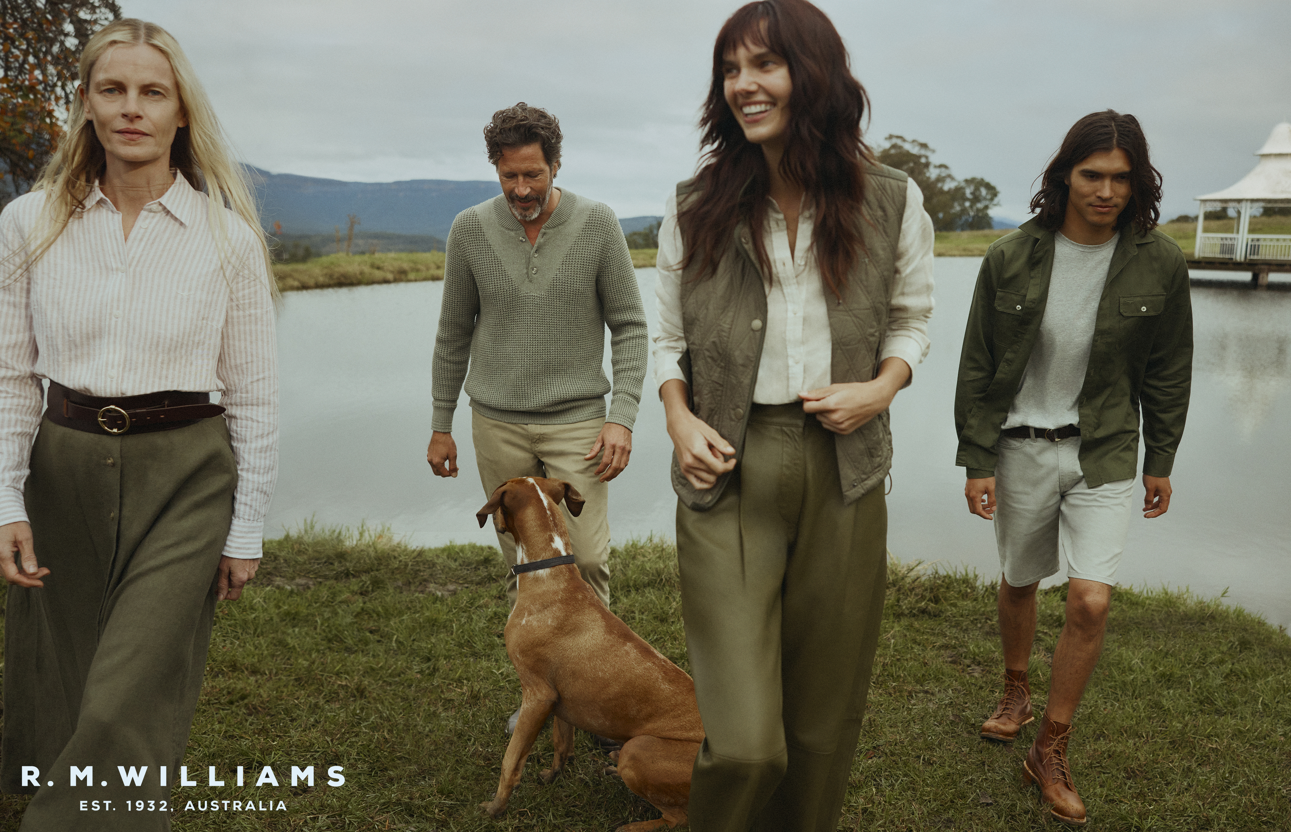 Group of four people and a dog are gathered in a grassy field near a pond, with a mountain range visible in the background on an overcast day.