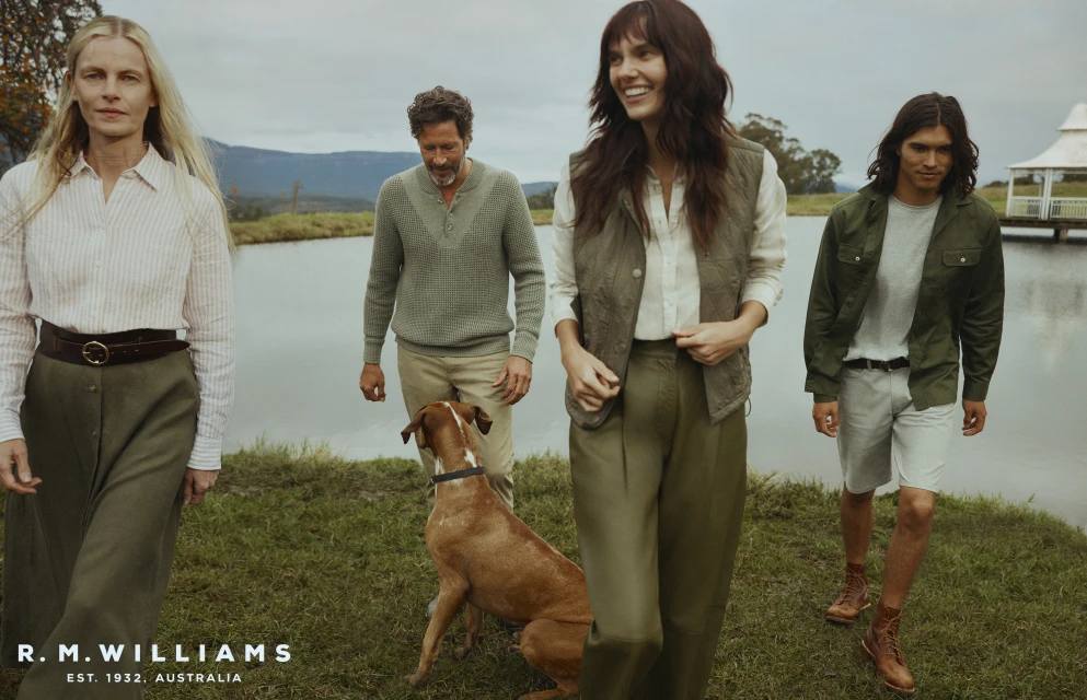 Group of four people and a dog are gathered in a grassy field near a pond, with a mountain range visible in the background on an overcast day.