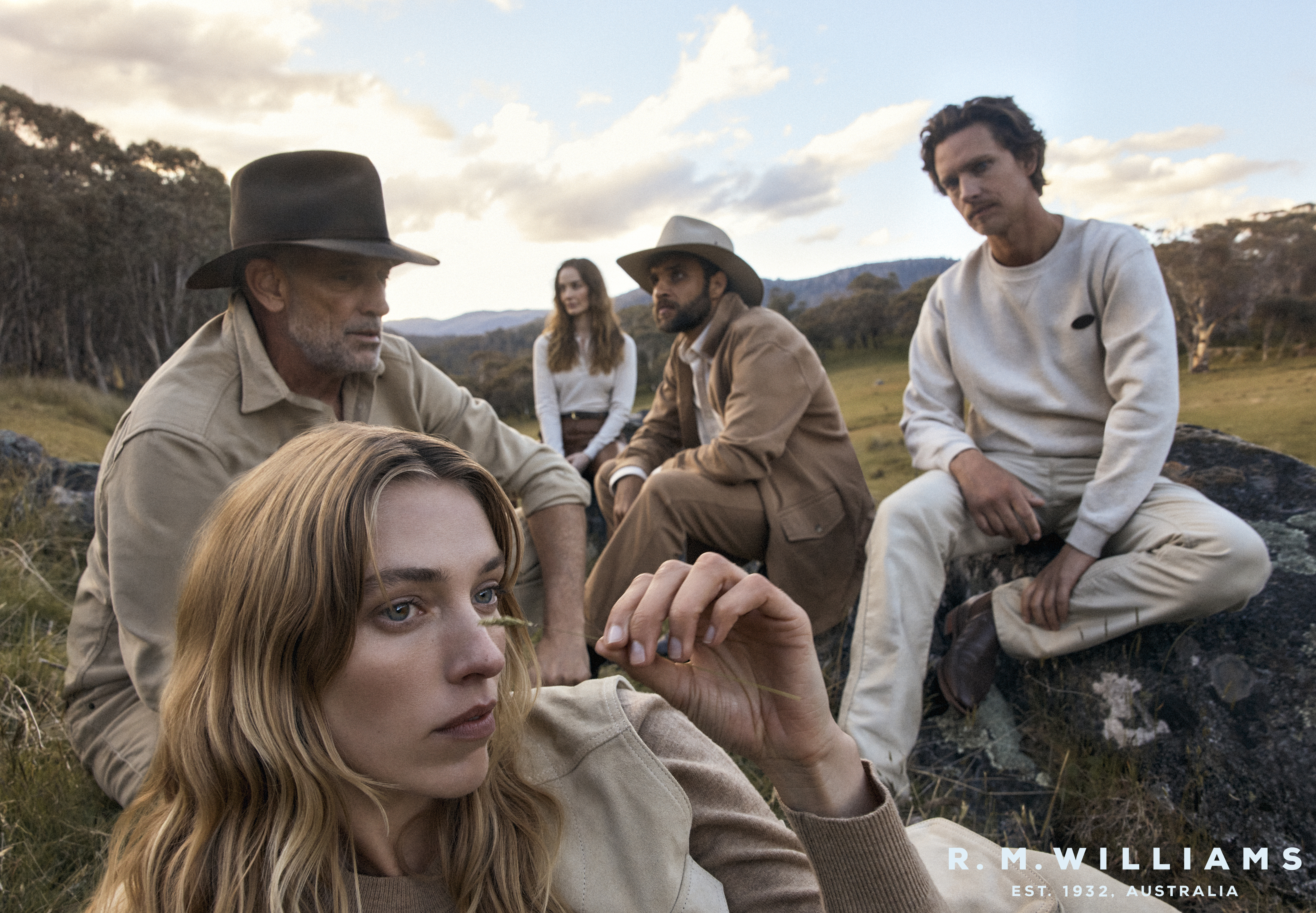 Five people in outdoor apparel sitting on rocks in a field surrounded by grassy hills and trees, under a cloudy sky, wearing hats.