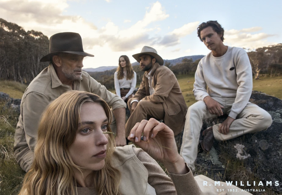 Five people in outdoor apparel sitting on rocks in a field surrounded by grassy hills and trees, under a cloudy sky, wearing hats.