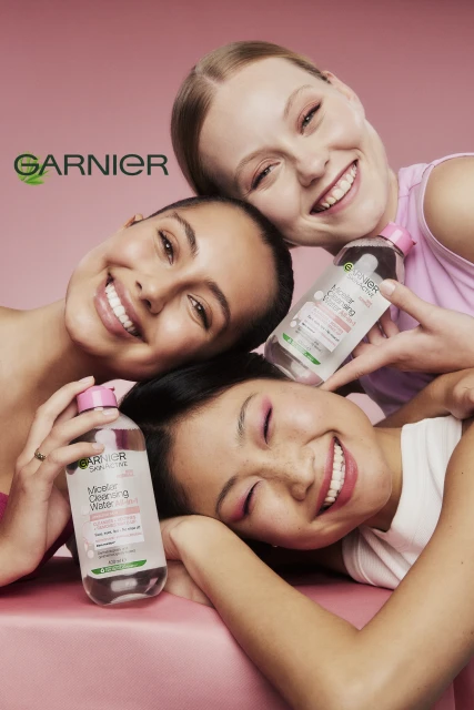 Three smiling young women pose with Garnier Micellar Cleansing Water bottles in a close-up shot against a solid pink background.