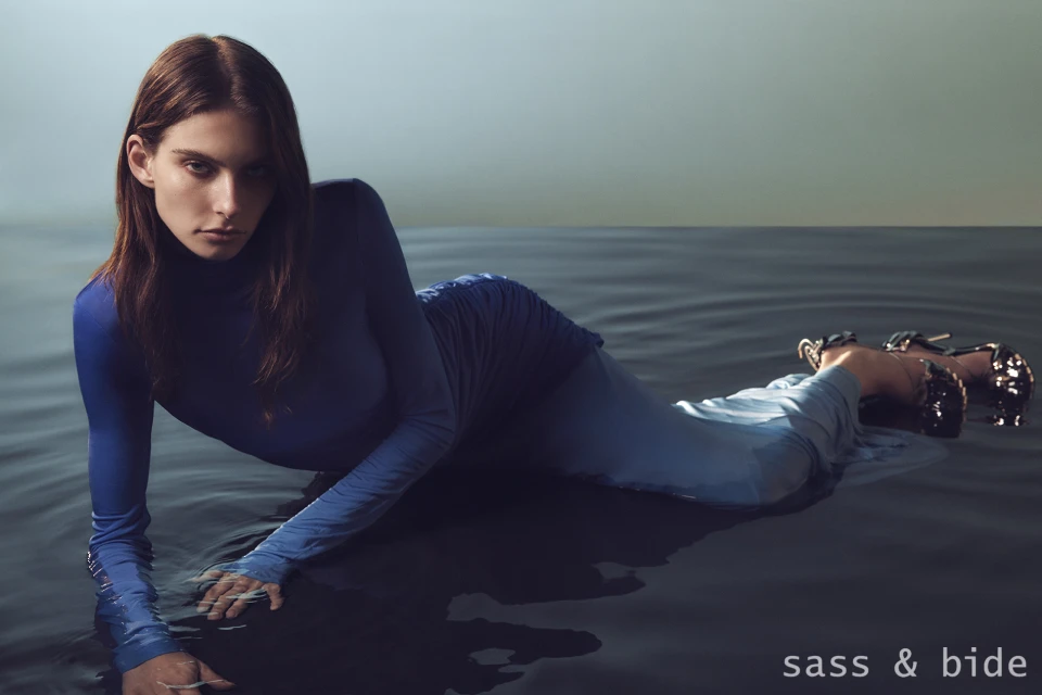Fashion shot of a woman in a blue dress lying in water, gazing at the camera with a serene expression, showcasing style and elegance.
