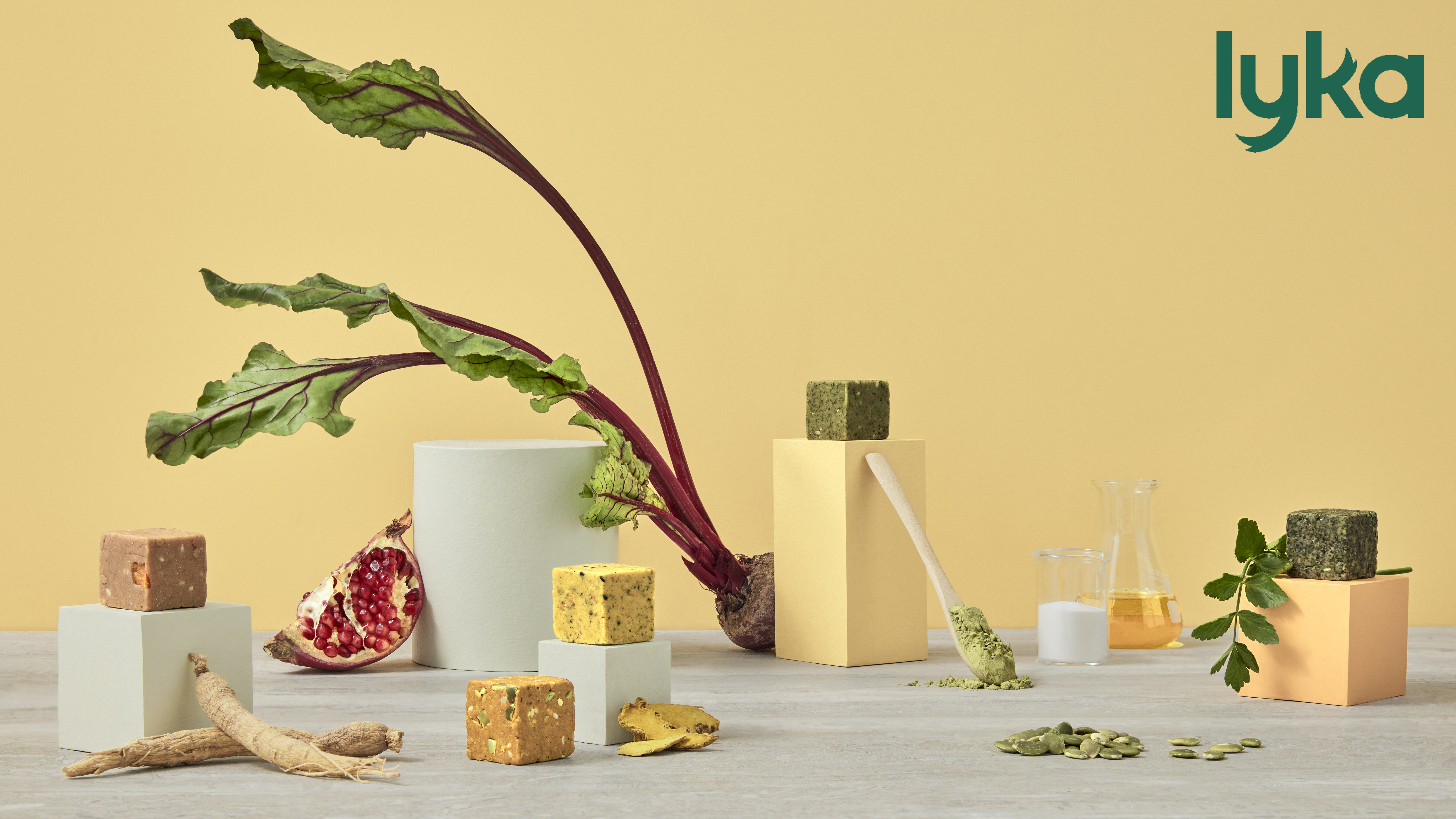 Still life featuring beet root with leaves, pomegranate half, cubes on pedestals, ginseng root, and glassware against yellow backdrop.