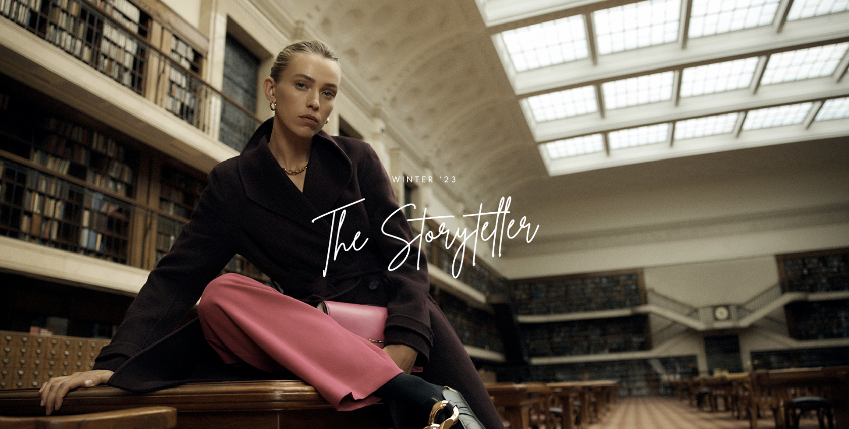 Indoor shot of a woman in a dark coat and pink trousers, sitting on a table in a library setting with bookshelves.