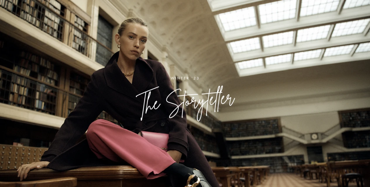 Indoor shot of a woman in a dark coat and pink trousers, sitting on a table in a library setting with bookshelves.