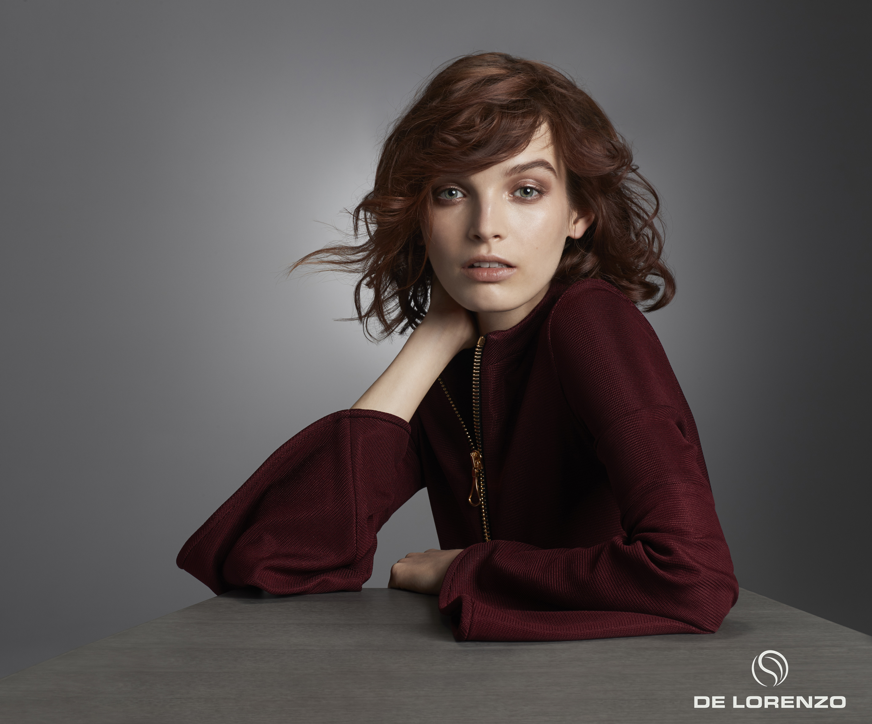 Portrait of a young woman with auburn wavy hair, wearing a burgundy top, posing against a gradient grey background, fashion shot.