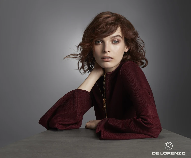 Portrait of a young woman with auburn wavy hair, wearing a burgundy top, posing against a gradient grey background, fashion shot.