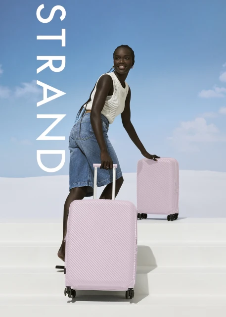 A smiling African woman in denim shorts and a top pulls two pink rolling suitcases outdoors against a sky backdrop.