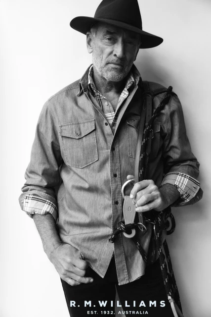 Portrait of an adult man wearing a hat and denim shirt, holding a metal clip on a rope, against a neutral background in black and white.