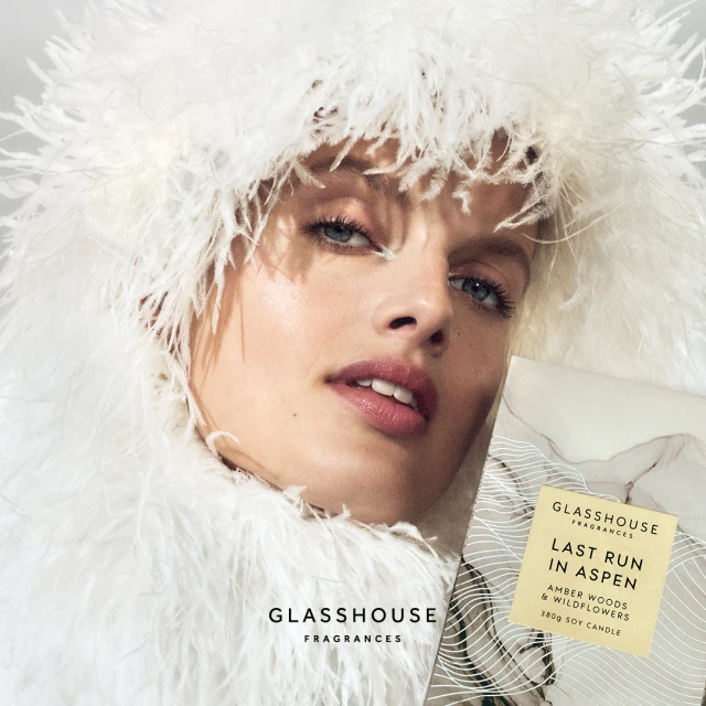 Close-up of a fair-skinned adult woman with light eyes in white feathered clothing, with Glasshouse Fragrances candle and branding.
