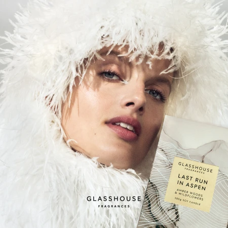 Close-up of a fair-skinned adult woman with light eyes in white feathered clothing, with Glasshouse Fragrances candle and branding.