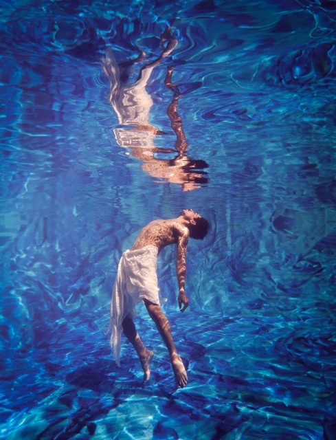 Underwater view of an adult male with bare torso wearing a white wrap, floating in blue water with light reflections on the surface.