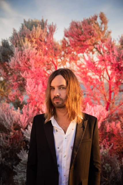 Portrait of adult male with long brown hair, wearing a black blazer and patterned white shirt, posing outdoors in front of pink trees.