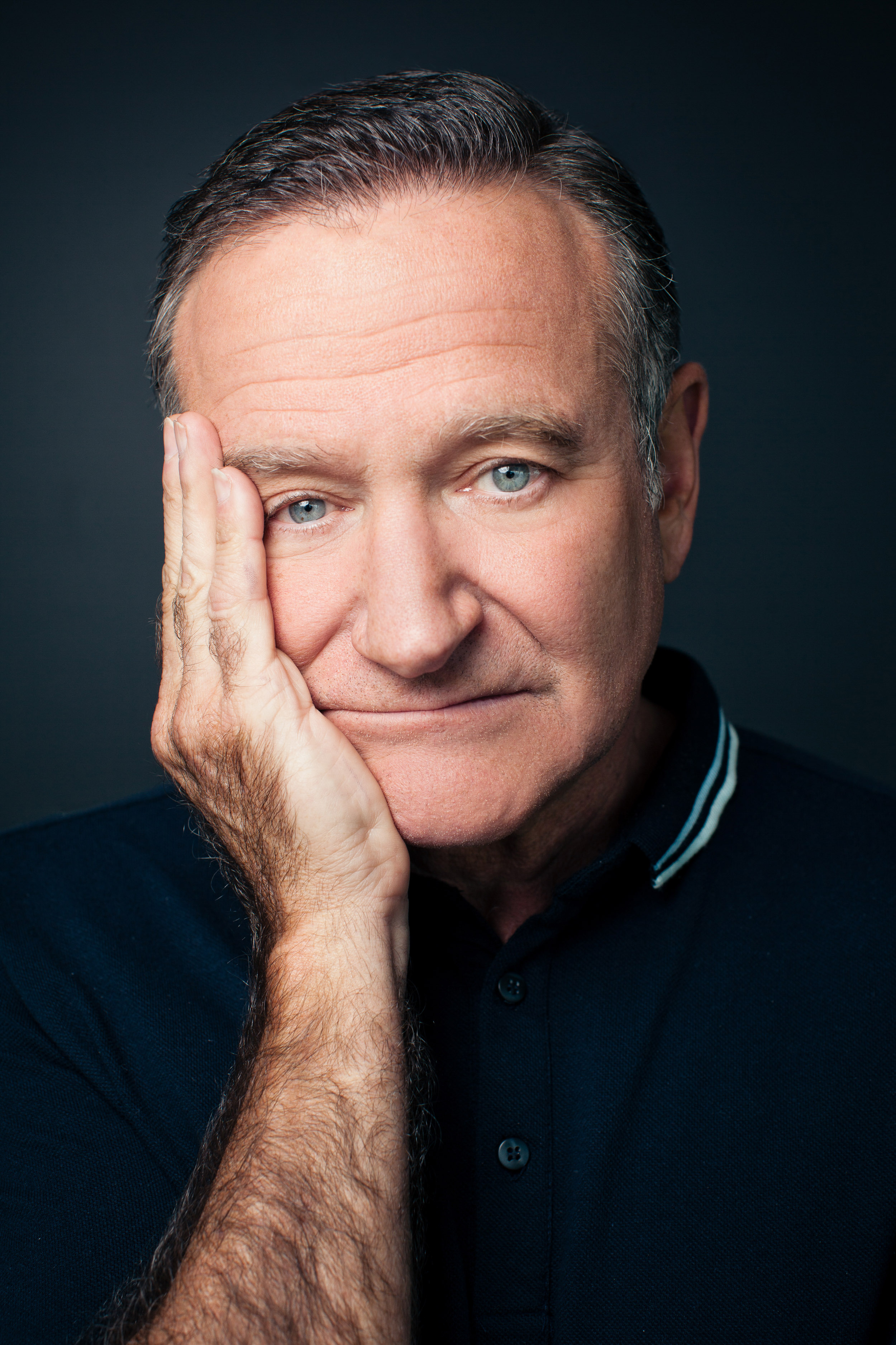 Close-up portrait of Robin Williams with his face resting on his hand, wearing a dark blue polo shirt against a dark background.