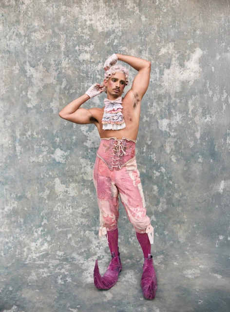 Man in rococo-inspired fashion with pink and white ruffled shirt, corset, bloomers, and shoes, posing against a textured grey background.