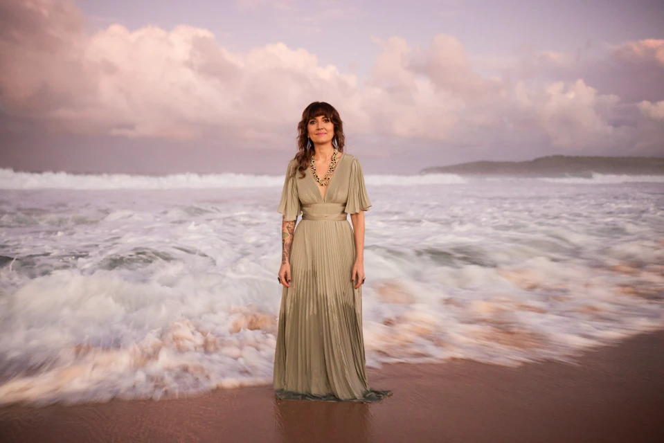 Woman in flowing dress standing on beach with ocean waves and pink sky at dusk, creating a serene and picturesque scene.