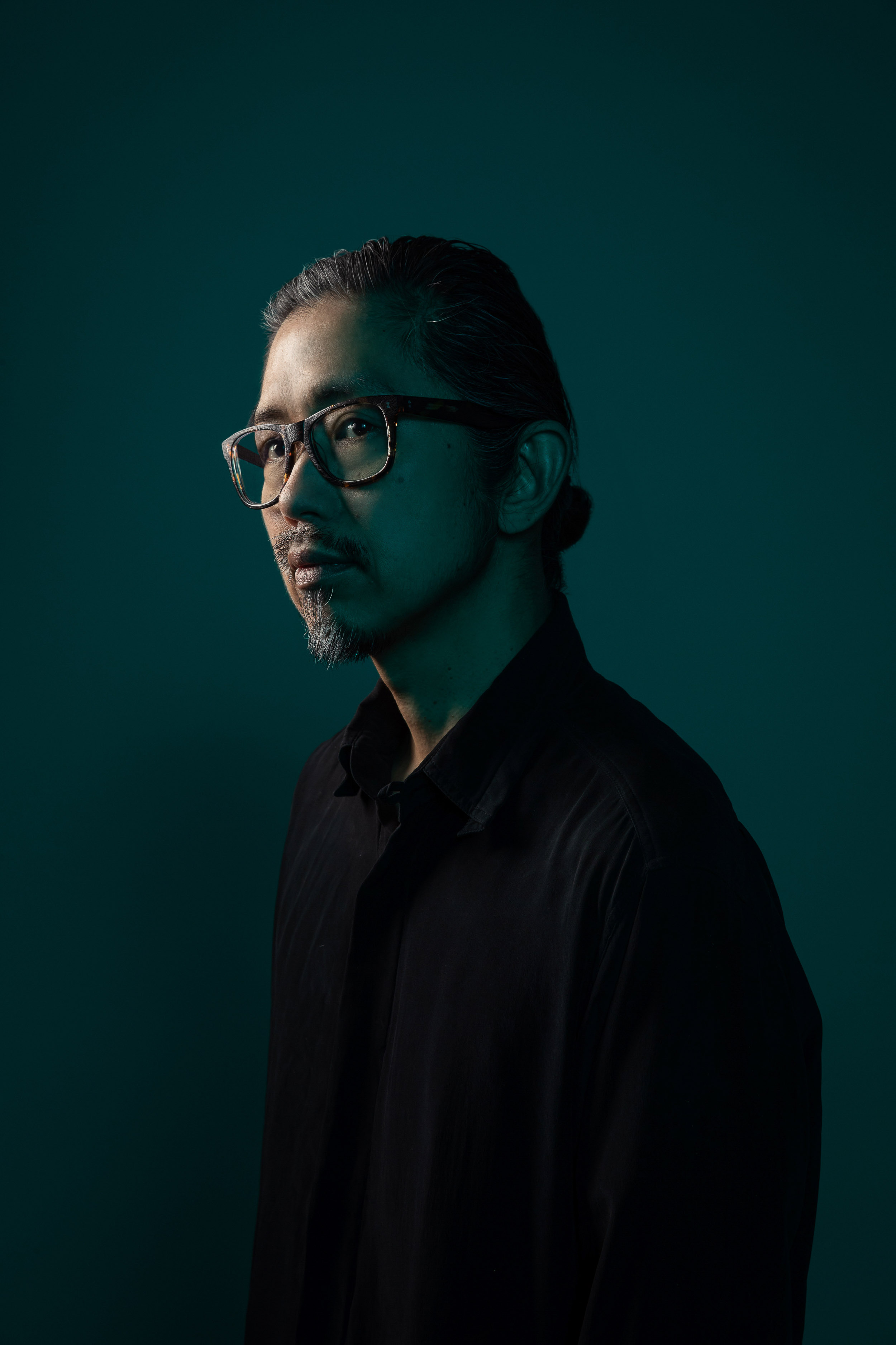 Portrait of an Asian man wearing glasses and a black shirt in a studio setting with teal lighting and a neutral expression.