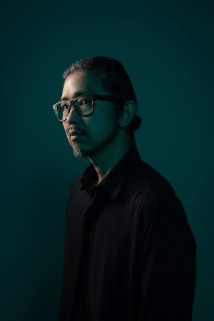 Portrait of an Asian man wearing glasses and a black shirt in a studio setting with teal lighting and a neutral expression.