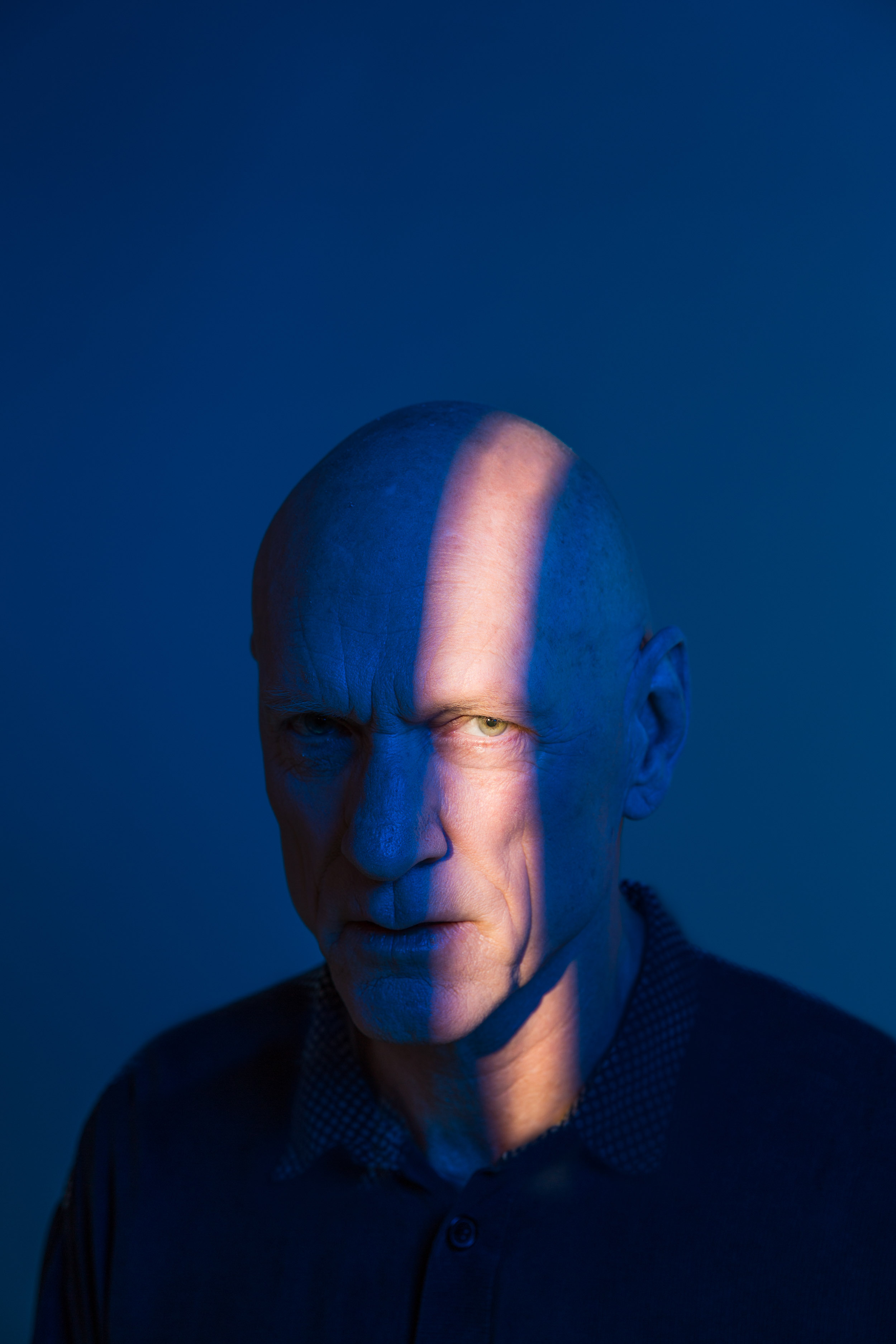 Portrait of a bald adult man with a serious expression, strongly lit with blue and orange hues in a studio setting.