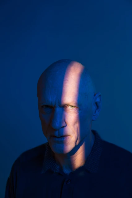 Portrait of a bald adult man with a serious expression, strongly lit with blue and orange hues in a studio setting.