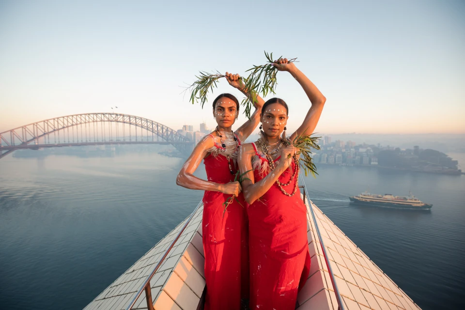Two Indigenous Australian women in red dresses pose with foliage on a rooftop overlooking the Sydney Opera House, bridge, and harbor.