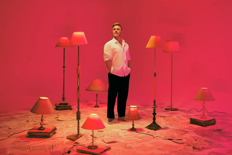 Man in white shirt and black pants stands in a room filled with lamps and sheet music under vibrant pink lighting, creating a surreal scene.