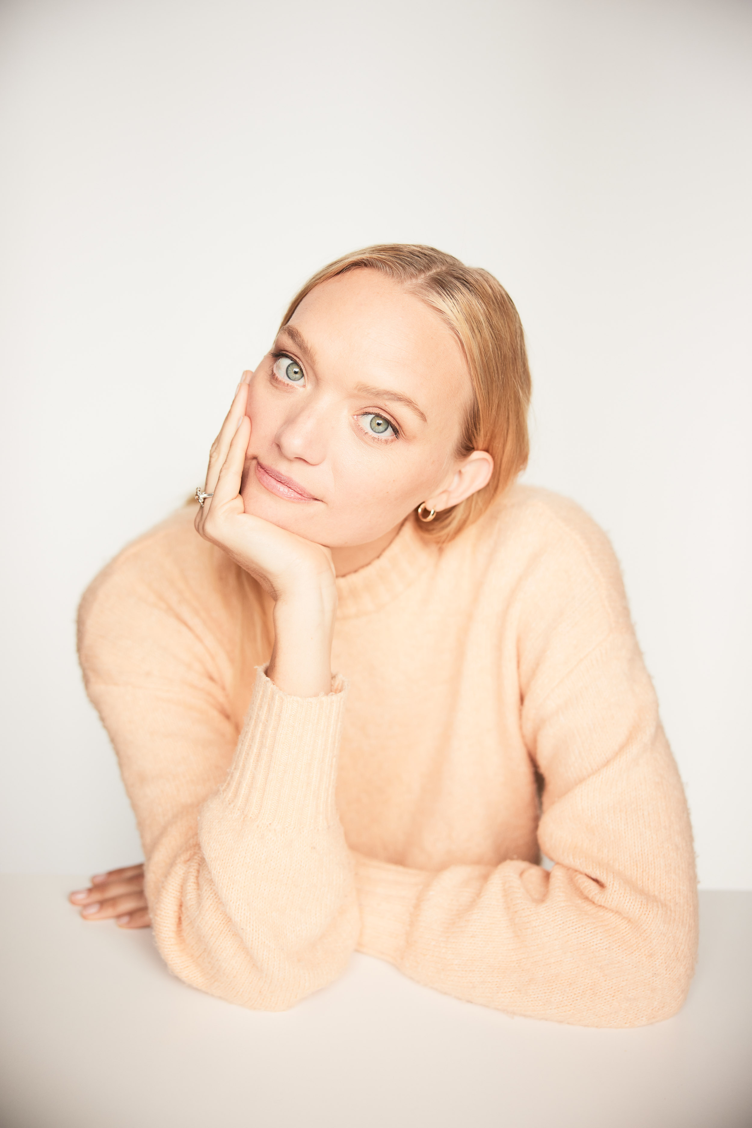 Portrait of a blonde woman with her hand on her face. She's wearing a peach sweater and is resting her elbows on a white table.