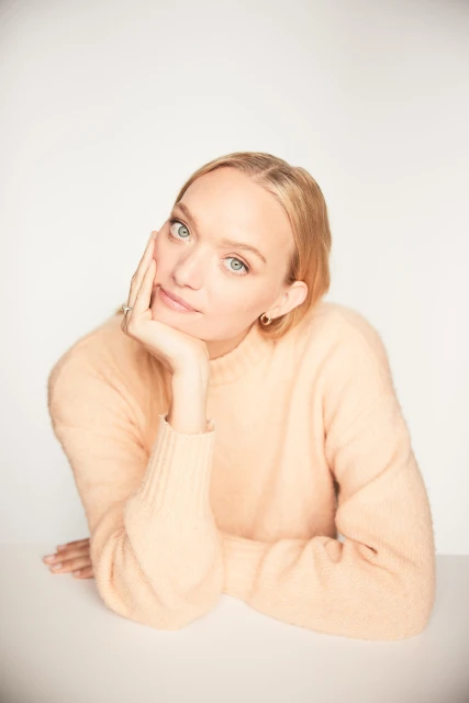 Portrait of a blonde woman with her hand on her face. She's wearing a peach sweater and is resting her elbows on a white table.