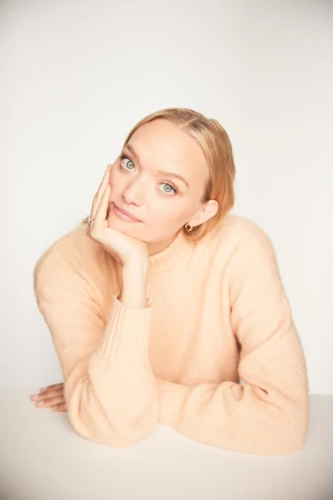 Portrait of a blonde woman with her hand on her face. She's wearing a peach sweater and is resting her elbows on a white table.