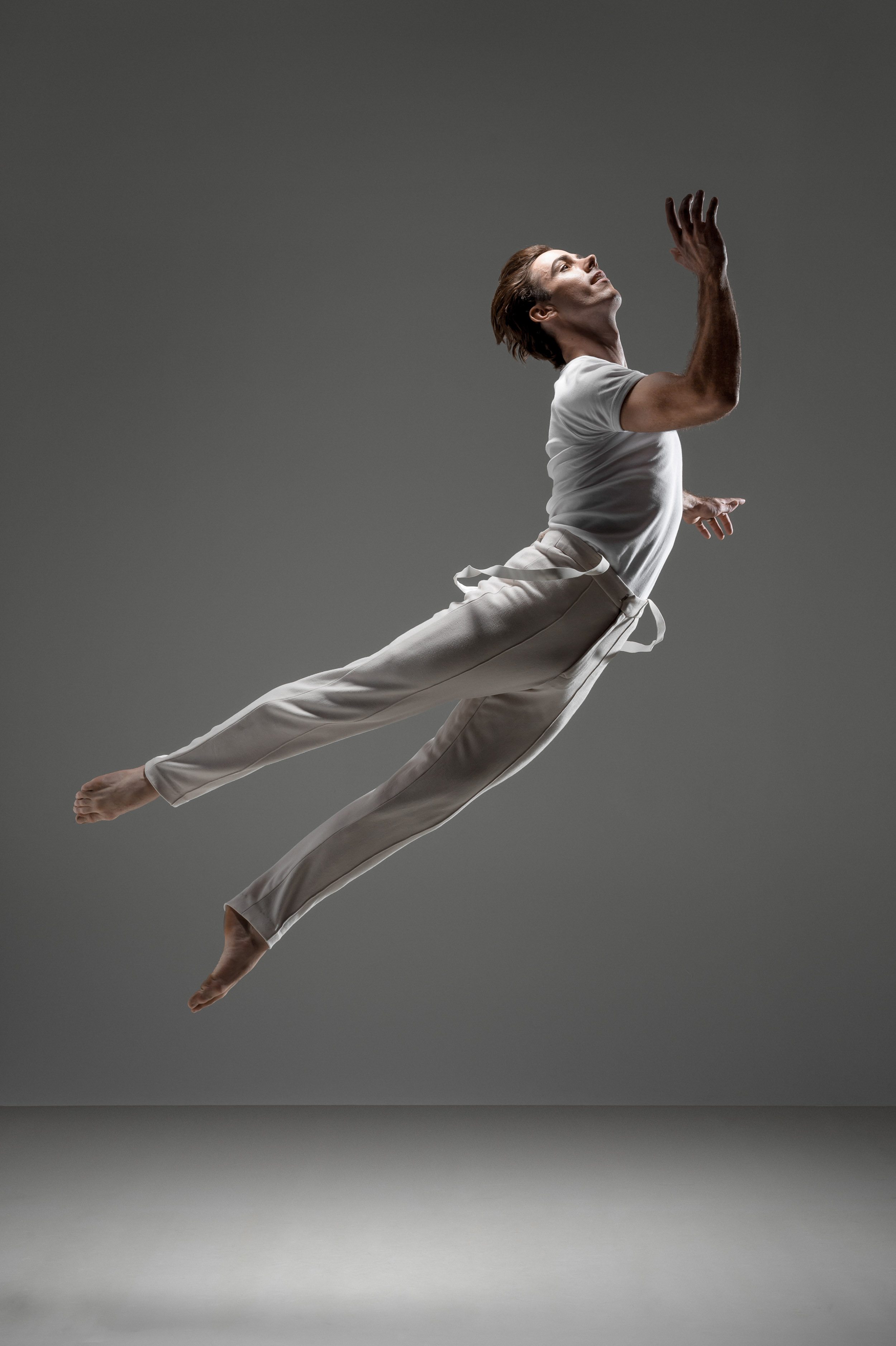 Male ballet dancer in white t-shirt and pants leaping in the air with outstretched arm against a grey gradient background in a photo studio.