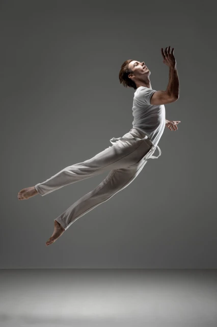 Male ballet dancer in white t-shirt and pants leaping in the air with outstretched arm against a grey gradient background in a photo studio.