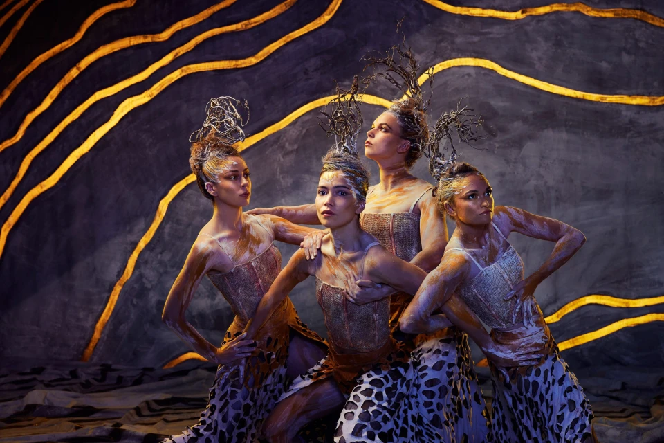 Four female dancers in ornate costumes and makeup pose in front of a stylized backdrop with gold accents for a theatrical performance.
