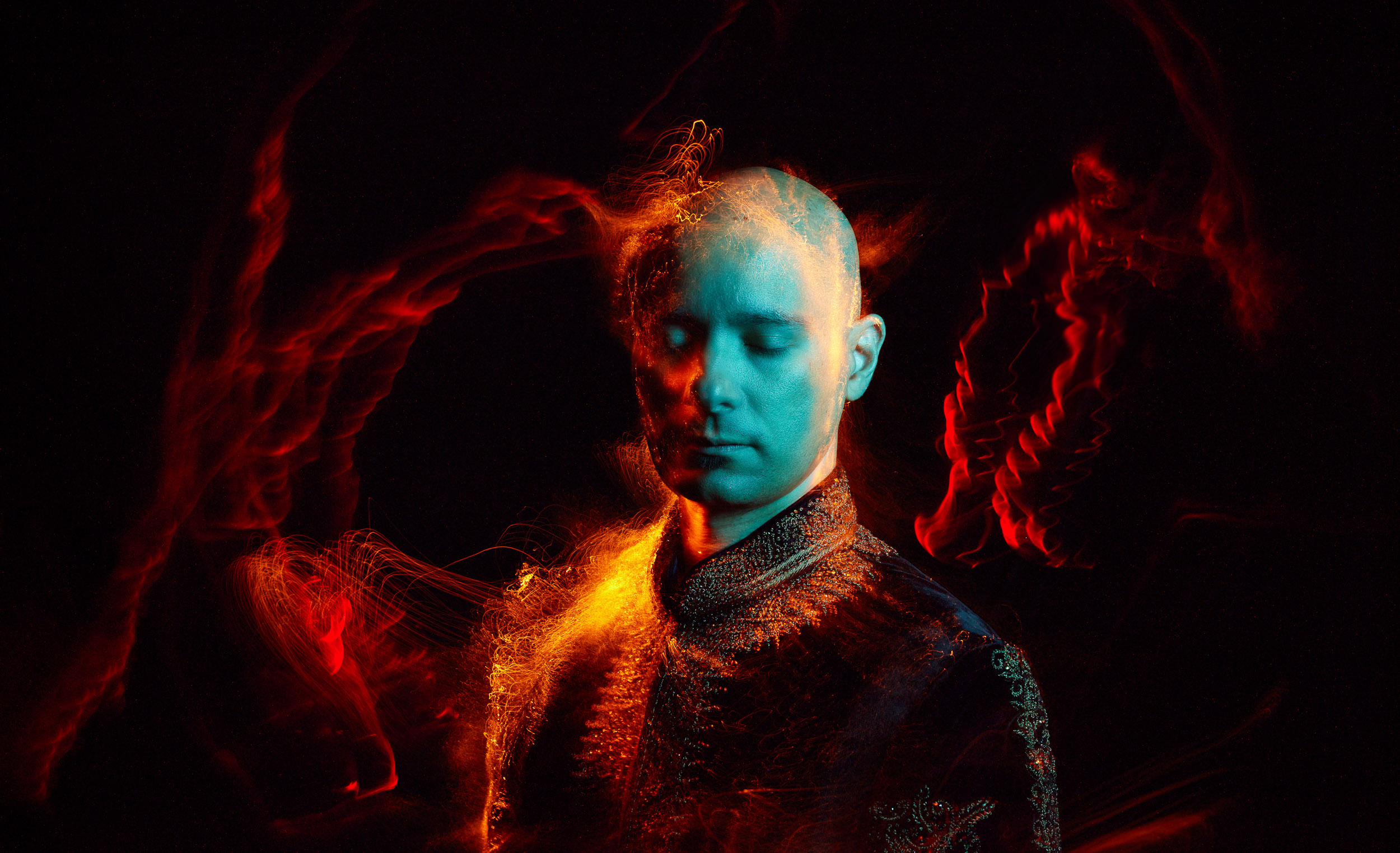 Portrait of an adult male with a shaved head wearing a patterned jacket against a black background with colorful light trails.