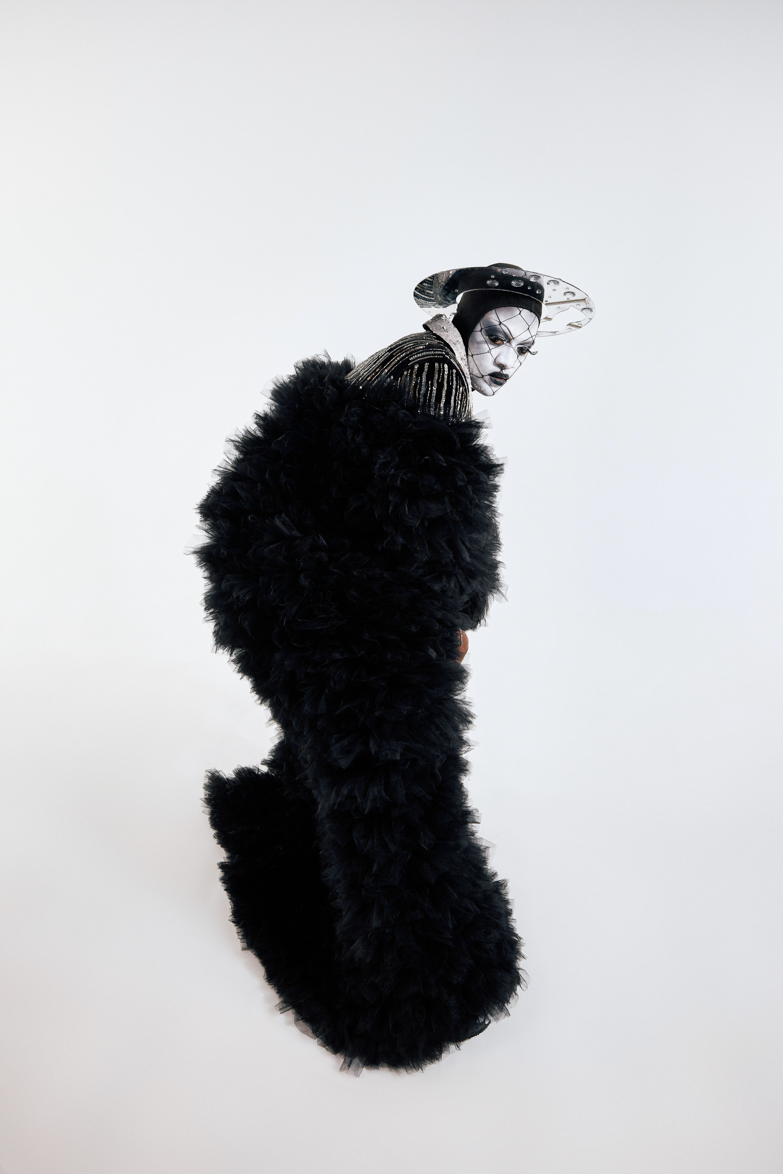 Person in avant-garde fashion with black fluffy coat, theatrical makeup, and stylish hat against a plain white backdrop in studio shot.