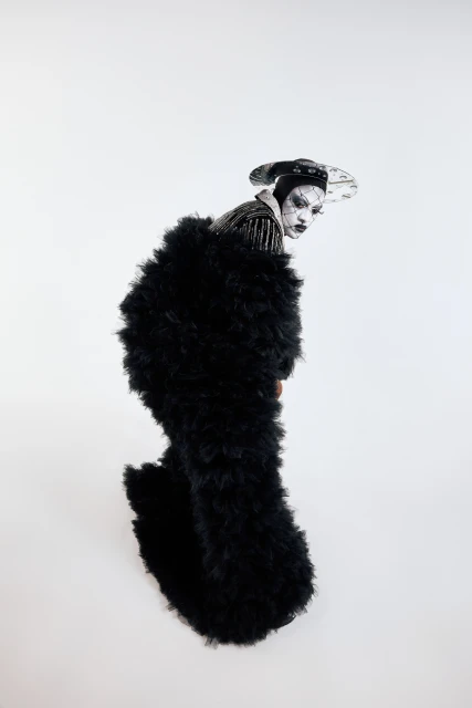 Person in avant-garde fashion with black fluffy coat, theatrical makeup, and stylish hat against a plain white backdrop in studio shot.