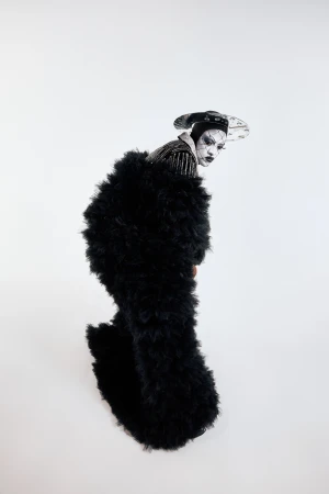 Person in avant-garde fashion with black fluffy coat, theatrical makeup, and stylish hat against a plain white backdrop in studio shot.