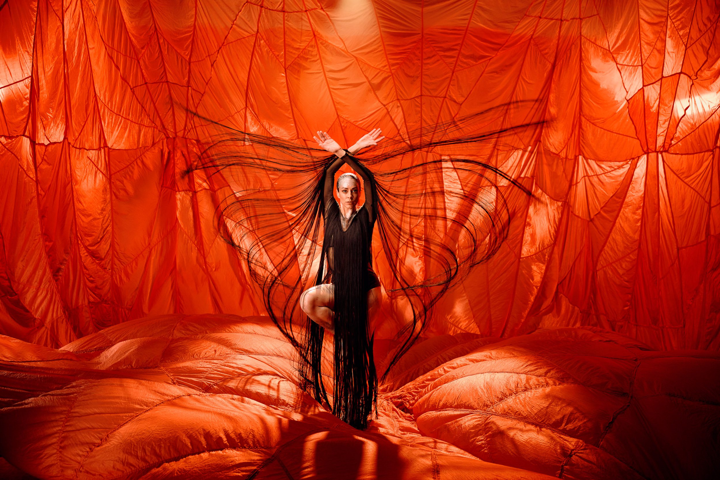 Artistic portrait of a woman in a black dress posing against an orange fabric backdrop with fringe details.