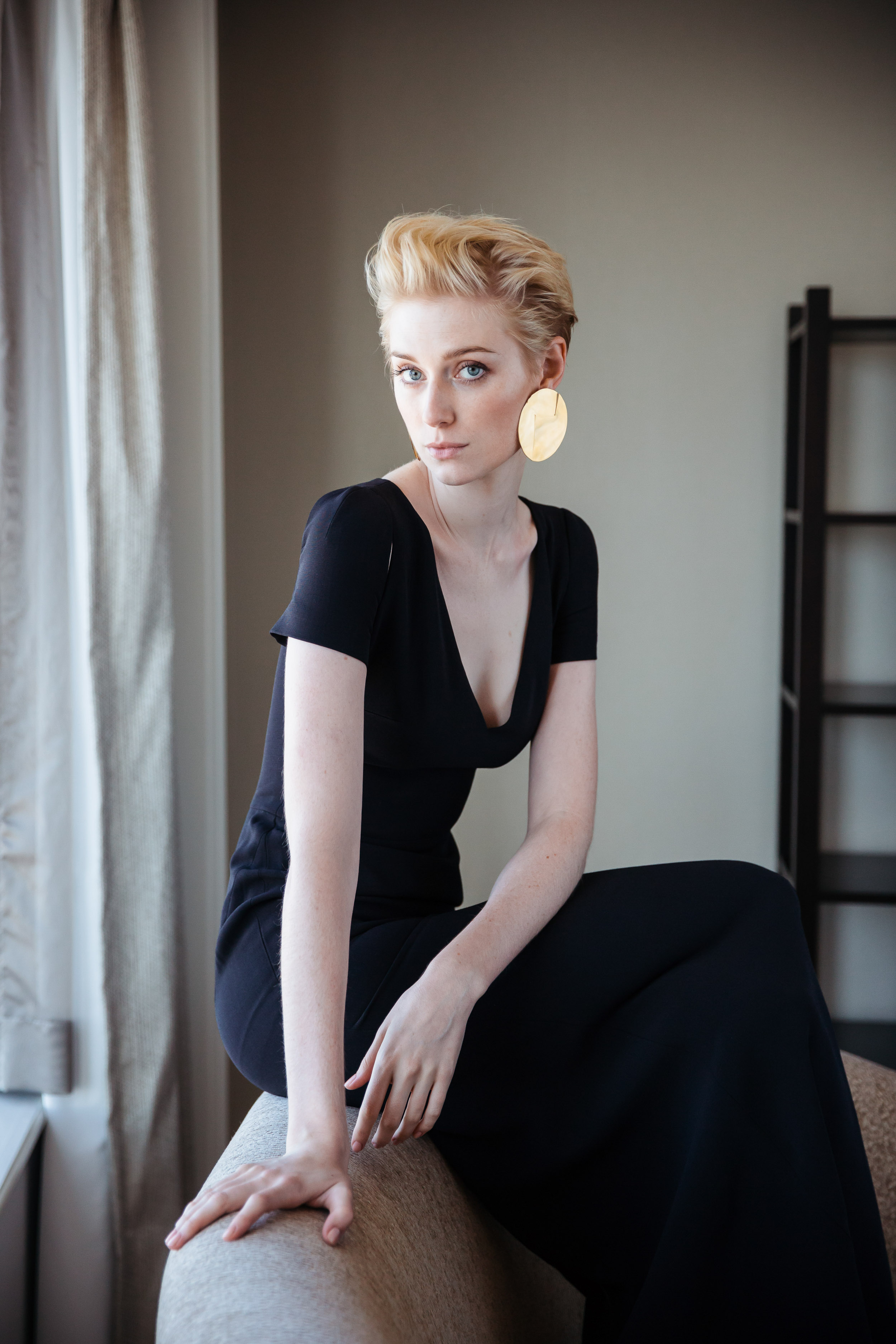 Elegant woman in a black dress seated indoors. The shot emphasizes fashion and portraiture with soft lighting and neutral tones.