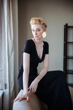 Blonde woman wearing a black dress and large gold earrings seated on a couch indoors by Daniel Boud