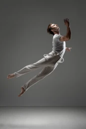 Male dancer captured mid-air in a dynamic leap, wearing a white t-shirt and trousers against a grey studio backdrop.