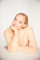 Portrait of a blonde woman wearing a peach sweater resting her chin on her hand against a white backdrop.