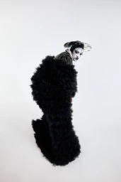 Model wearing a voluminous black ruffled coat and patterned hat with veil against a white background