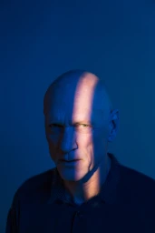 Portrait of a mature bald man in a dark polo shirt lit by a sharp vertical stripe of warm light against a blue background.