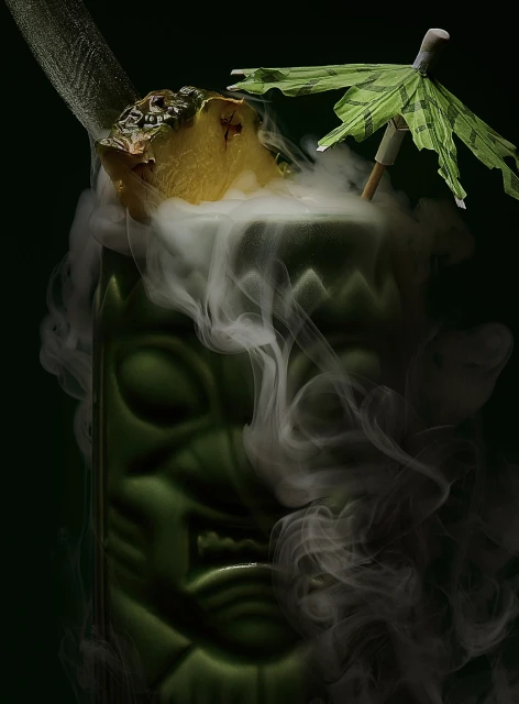 Exotic tiki cocktail in a green tiki-style glass with pineapple garnish, paper umbrella, and swirling smoke, set against a dark background.