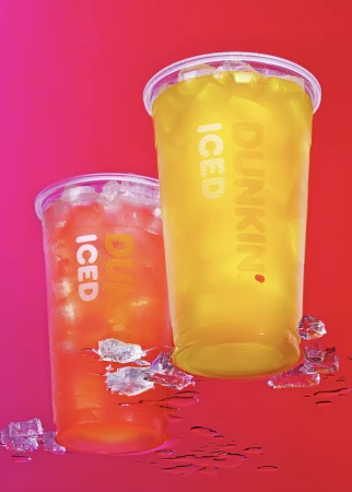 Two Dunkin' iced drinks in plastic cups with ice cubes, one yellow and one red-orange, on a vivid pink and red surface.