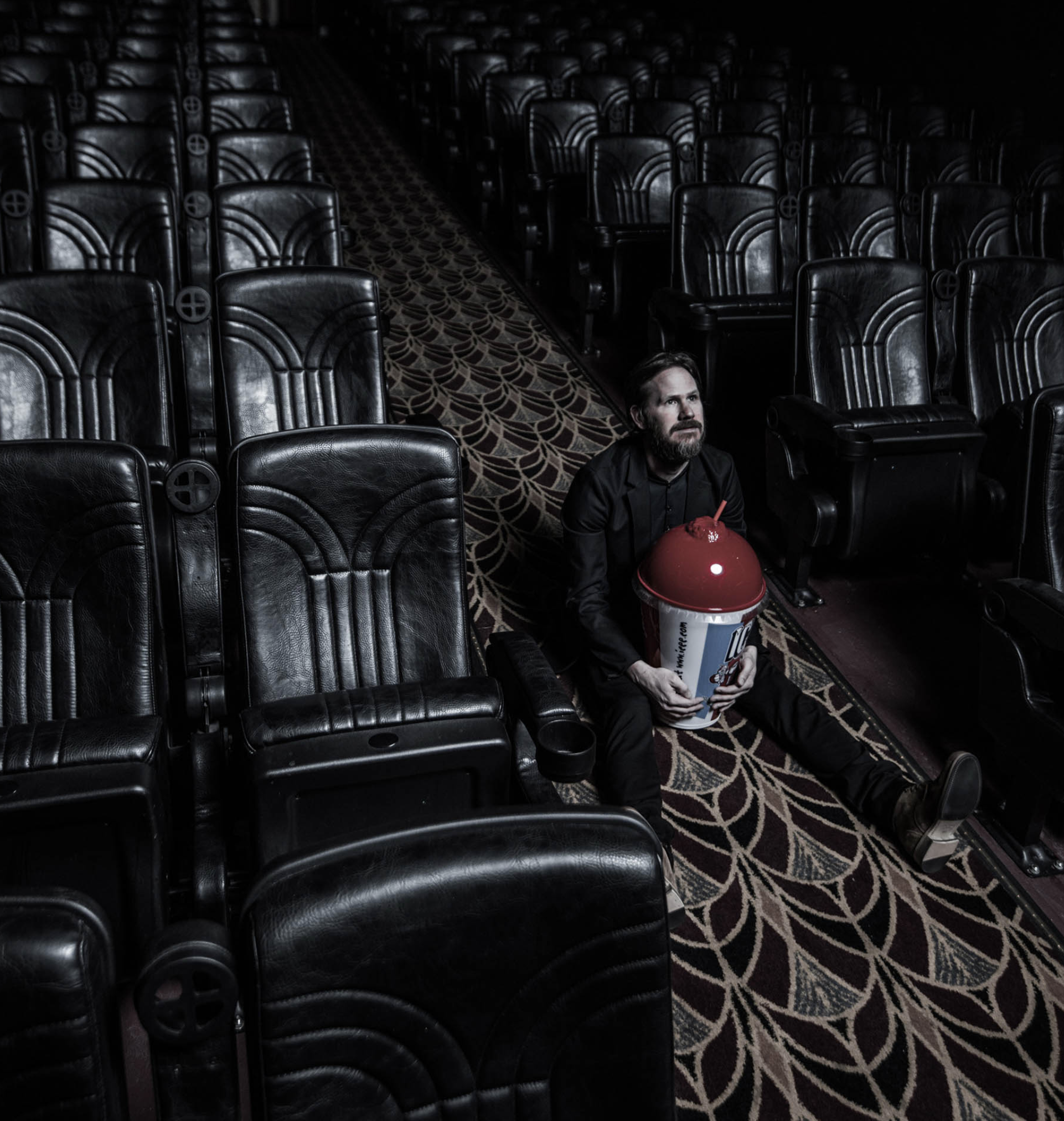 Man sits alone in a dark movie theatre, holding a large tub of popcorn, surrounded by empty seats, creating a solitary, cinematic scene.