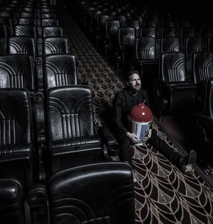 Man sits alone in a dark movie theatre, holding a large tub of popcorn, surrounded by empty seats, creating a solitary, cinematic scene.